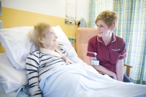 A female staff member in a raspberry tunic having a conversation with a smiling elderly female patient in a hospital bed in a ward.