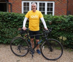 Mike Gwynn in a yellow QVH Charitable Fund T-shirt and bike shorts, posing with a bicycle in front of some bushes and a brick building.