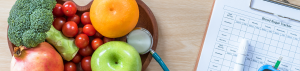 A wooden plate of healthy vegetables and fruit to the left and next to it on the right is a clipboard and pen
