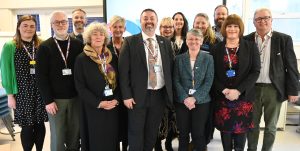 Members of the board of directors standing together in a group. They are all wearing lanyards with the progress pride flag on it, and matching badges. They are all smiling at the camera